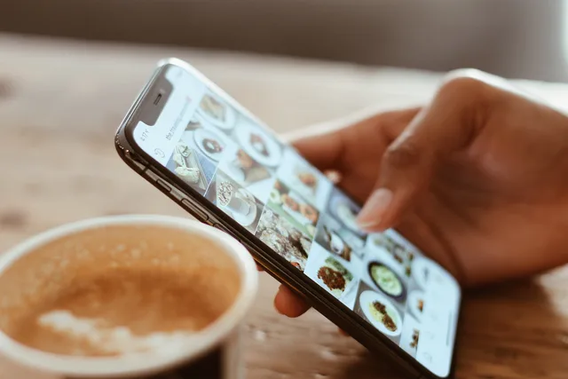 Hand browsing social media photos on a smartphone next to a cup of coffee indoors. Coordonner e-mails et réseaux sociaux en Suisse : rythme et contenus
